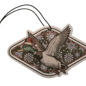 May include: A decorative air freshener featuring a flying duck wearing a Santa hat. The duck is depicted with detailed feathers and a green head. The background has a snowflake pattern. A black string is attached for hanging.