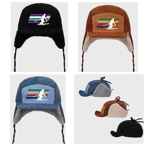 May include: A collection of corduroy trapper hats in black, brown, and blue. Each hat features a rectangular patch with a skier graphic and horizontal stripes. The hats have ear flaps and chin ties for a secure fit.