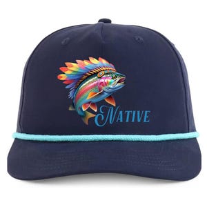 May include: Navy blue baseball cap featuring a colorful illustration of a rainbow trout wearing a Native American headdress. The word "NATIVE" is written in a script font below the fish. The cap has a light blue rope detail.