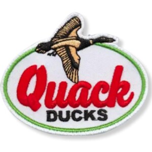 Könnte beinhalten: Ein weißer ovaler Patch mit grünem Rand. Der Patch zeigt das Wort "Quack" in roter Stickerei mit dem Wort "Ducks" in Schwarz darunter. Ein Cartoon-Ente fliegt über dem Text.