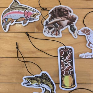 May include: Air fresheners with various outdoor themes. Designs include a rainbow trout, a hunting dog with a duck, a fish, and a shotgun shell. Each air freshener is attached to a black string for hanging.