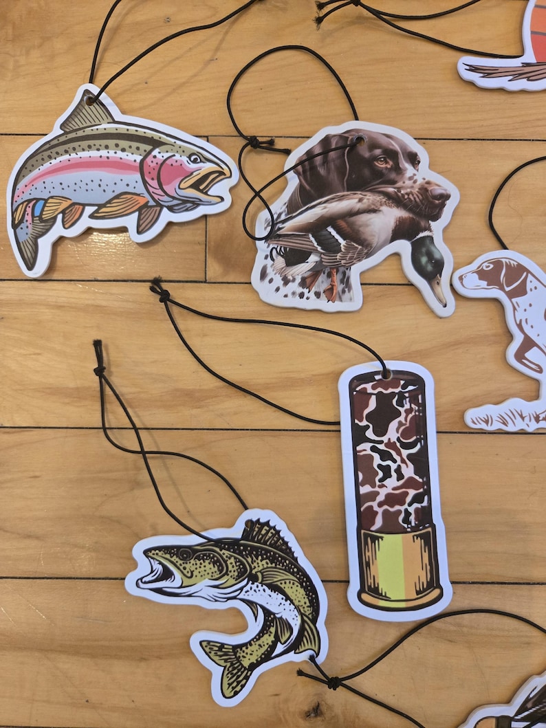 May include: Air fresheners with various outdoor themes. Designs include a rainbow trout, a hunting dog with a duck, a fish, and a shotgun shell. Each air freshener is attached to a black string for hanging.