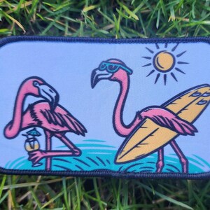 May include: A blue and white patch with two pink flamingos. One flamingo is holding a cocktail and the other is holding a surfboard. The sun is shining in the background.