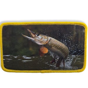 Puede incluir: Un parche rectangular con borde amarillo muestra un lucio saltando fuera del agua. El pez está detallado con colores naturales y gotas de agua, sugiriendo movimiento. El fondo es una escena de bosque borrosa.