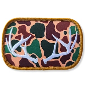 Pode incluir: Um remendo bordado branco em forma de chifre de veado sobre um fundo de camuflagem marrom, verde e laranja. O remendo tem uma borda costurada dourada.
