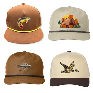 Pu&ograve; includere: Quattro cappellini con diversi disegni. Un cappellino marrone con un pesce, un cappellino beige con un armadillo, un cappellino marrone con una mosca da pesca e un cappellino con un'anatra mimetica.