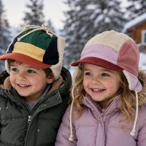 May include: Two corduroy trapper hats with ear flaps and chin ties. The left hat features a color-blocked pattern of green, yellow, navy, and a rust-colored brim. The right hat features a color-blocked pattern of cream, light pink, and a maroon brim.