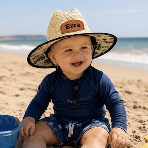 Peut inclure: Un chapeau en paille à large bord avec une bordure noire et un dessous à motifs noirs et blancs. Un écusson en cuir marron sur le devant du chapeau porte le nom Ezra gravé en lettres noires. Le chapeau est muni d'une mentonnière noire.