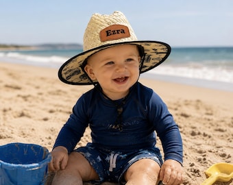 May include: A straw hat with a wide brim featuring a black trim and a black and white patterned underside. A brown leather patch on the front of the hat is embossed with the name Ezra in black lettering. The hat includes a black chin strap.