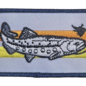 May include: Embroidered patch depicting a fish with a fishing hook. The fish is white with black spots, set against a light blue background with orange and yellow stripes. The patch has a dark blue border.