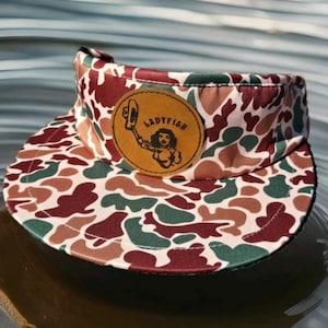 May include: A camouflage patterned visor with a brown leather patch that says "Ladyfish" in black lettering. The patch features a cartoon image of a woman wearing a cowboy hat and holding a fishing rod.