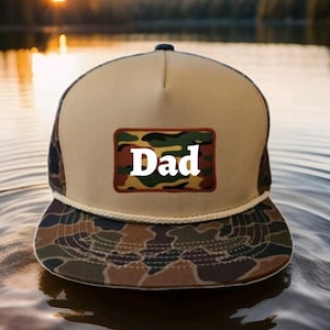 Könnte beinhalten: Eine beige und camouflagefarbene Trucker-Kappe mit weißem Kordelbesatz. Die Kappe hat einen Camouflage-Aufnäher mit dem Wort "Dad" in weißen Buchstaben.