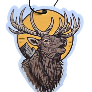 May include: An air freshener with a brown elk design. The elk has large antlers and is set against a yellow sun and mountain scene. The air freshener has a light blue border and a black string for hanging.