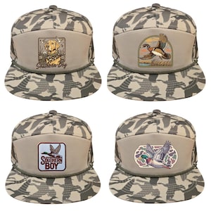 May include: Four camouflage trucker hats with embroidered patches. The patches feature a yellow Labrador, a flying duck, and the text "Southern Boy". The hats have a beige front panel and a green rope detail.