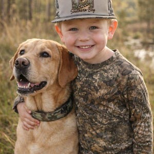 May include: A young child wearing a camouflage hat and shirt, hugging a golden Labrador Retriever. The hat features a graphic of a dog and the text "Southern Boy". The dog wears a camouflage collar. The child is smiling.