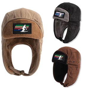 Könnte beinhalten: Drei Trappermützen aus Cord mit Ohrenklappen. Die Mützen sind braun, schwarz und beige. Jede Mütze hat einen Aufnäher mit einem Skifahrer darauf. Die Aufnäher haben ein Regenbogenstreifen-Design.