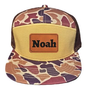 Op de afbeelding: Een bruine en beige camouflage trucker cap met een beige klep en een bruin mesh achterkant. De pet heeft een bruin lederen patch met de naam "Noah" in zwarte letters.