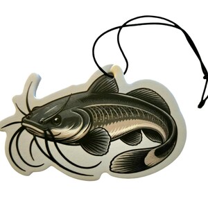 May include: A decorative air freshener shaped like a catfish. The air freshener features a detailed illustration of the fish in shades of gray and beige, with long whiskers and a black string for hanging.