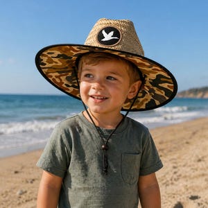 May include: A straw sun hat with a camouflage brim and a bird patch. The hat is worn by a child on a beach. The child is wearing a green t-shirt and a black string around the neck.