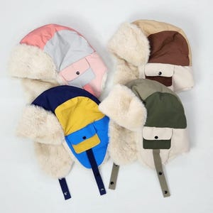 May include: Four colorful winter hats with ear flaps. The hats feature a mix of colors, including pink, blue, yellow, brown, and green, with faux fur trim. Each hat has a small pocket and a strap with a button closure.