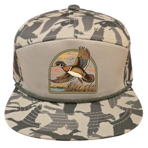 Puede incluir: Gorra de ala plana con un patrón de camuflaje y un parche con un pato volando sobre un paisaje de pantano. El pato está representado con plumas marrones, blancas y azules, sobre un cielo de atardecer. La gorra tiene un detalle de cuerda.