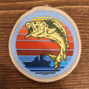 Puede incluir: Parche bordado con un pez saltando frente a un diseño de puesta de sol con rayas naranjas, rojas y azules. Una silueta de un barco con dos figuras pescando está en la parte inferior. El parche tiene un borde azul claro y un borde beige.