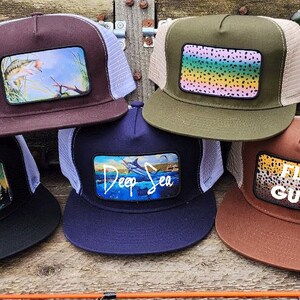 May include: Five different trucker hats with various fishing-themed patches. The patches feature a fish jumping out of the water, a trout, a marlin, a fly fishing pattern, and the text "Fly Guy".