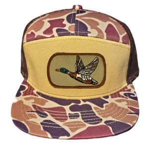 Puede incluir: Una gorra de camionero de camuflaje marrón y beige con un panel frontal beige y una malla trasera marrón. El panel frontal presenta un parche ovalado beige con una imagen bordada marrón de un pato volando.