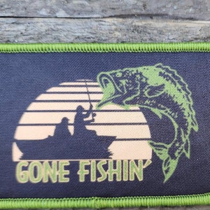 Puede incluir: Parche bordado verde y negro con una silueta de una persona pescando en un bote con un pez grande saltando fuera del agua. El texto "GONE FISHIN'" está debajo de la imagen.