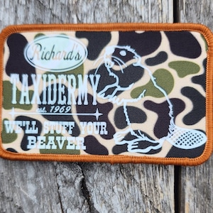 Peut inclure: Un patch en tissu camouflage brun et vert avec un contour blanc d'un castor et le texte "Richard's Taxidermy est. 1969 We'll Stuff Your Beaver".
