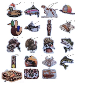 May include: A collection of air fresheners with hunting and fishing themes. Designs include a duck wearing a Santa hat, a trout, a hunting dog, a pheasant, a duck with peace sign, a fly, a duck call, a flying bird, a turkey, a wood duck, a bass, a deer, and a fish. Each has a string for hanging.