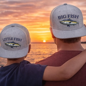 May include: Two gray trucker hats with fish illustrations and text. One hat says "BIG FISH" and the other says "LITTLE FISH". The hats are worn by two people, with a sunset over the ocean in the background.