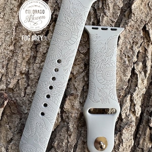 May include: A fog grey watch strap with a detailed dragon design. The strap is made of a soft material and features the text "Colorado Maven" and "Fog Grey". The watch strap is shown on a textured wood background.