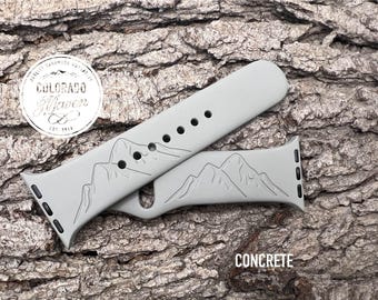 May include: A concrete-colored silicone watch band featuring an engraved mountain range design. The band includes a series of adjustment holes and black attachment hardware. A circular logo with the text Colorado Maven Est. 2019 is visible.