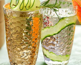 May include: Two textured acrylic glasses filled with a clear liquid and garnished with cucumber and orange slices. One glass has a golden hue, the other is clear. The glasses are part of a set of eight, each holding 23 oz.