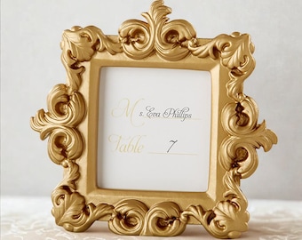 May include: A gold-colored, ornate picture frame with a square opening. The frame has elaborate scrollwork and a white insert with the text "Ms. Eva Phillips" and "Table 7". Ideal for weddings or special events.