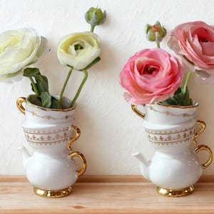 May include: Two white ceramic vases, each shaped like a teapot and teacup stacked, with gold accents and floral arrangements. One vase holds white flowers, the other pink. The vases sit on a wooden surface.