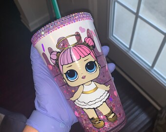 May include: A clear plastic tumbler with a pink glitter base and a pink and white cartoon doll design. The tumbler has a white rim with a row of rhinestones and a green straw.