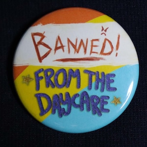 Puede incluir: Un botón redondo con fondo blanco y el texto "Banned! From the Daycare" en letras rojas y moradas. El botón tiene un borde amarillo, naranja y azul.