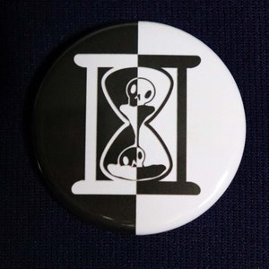 Puede incluir: Un botón blanco y negro con un diseño de reloj de arena. El reloj de arena está dividido en dos, con una calavera en la parte superior e inferior. El reloj de arena está rodeado por un marco blanco y negro.