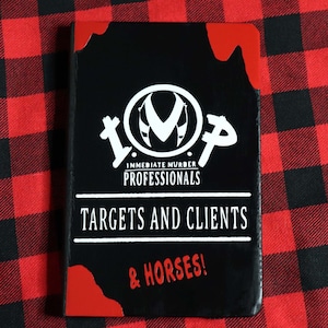May include: A black notebook with a white logo that says "IMP - Immediate Murder Professionals" and the text "Targets and Clients & Horses!" in white lettering. The notebook has red blood splatter on the cover.
