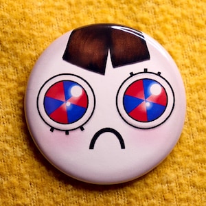 May include: A circular badge featuring a cartoon face. The eyes are a mix of red, blue, and white, with black detailing. The badge has a brown hair design and a curved black mouth. The background is yellow.