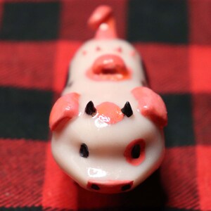 K&ouml;nnte beinhalten: Eine kleine, wei&szlig;e Keramik-Schweinefigur mit rosa Ohren, einer rosa Schnauze und schwarzen Augen. Das Schwein hat eine gl&auml;nzende Oberfl&auml;che und sitzt auf einem rot-schwarz karierten Hintergrund.