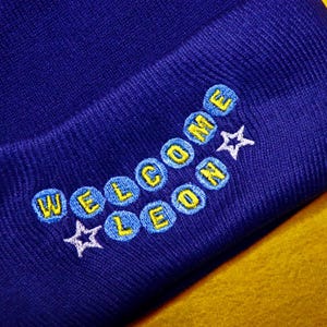May include: A close-up of a navy blue beanie hat with the embroidered text "WELCOME LEON" in yellow and blue, with white star accents. The hat is on a yellow surface.