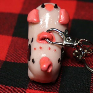 K&ouml;nnte beinhalten: Ein wei&szlig;er und schwarz gefleckter Schweine-Schl&uuml;sselanh&auml;nger mit rosa Ohren und einer rosa Schnauze. Das Schwein ist an einem silbernen Schl&uuml;sselanh&auml;nger aus Metall befestigt.