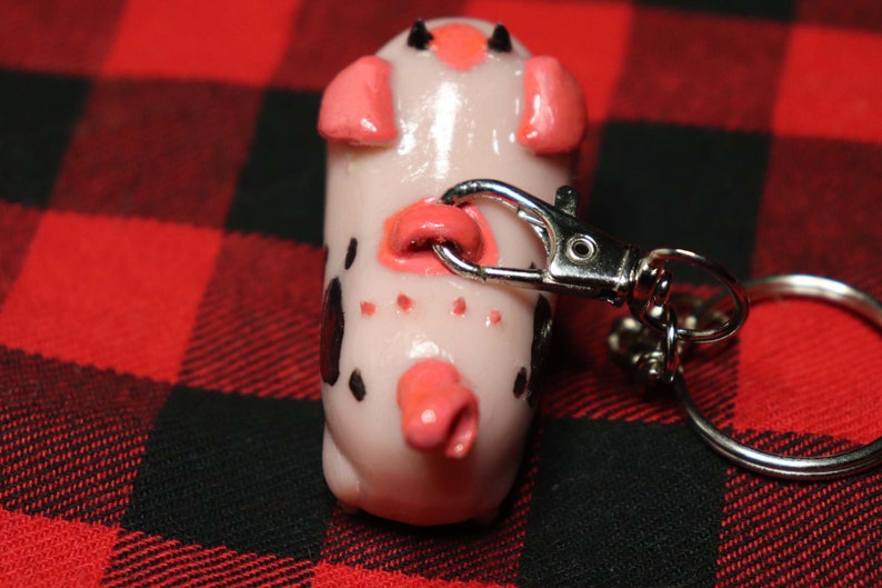 K&ouml;nnte beinhalten: Ein wei&szlig;er und schwarz gefleckter Schweine-Schl&uuml;sselanh&auml;nger mit rosa Ohren und einer rosa Schnauze. Das Schwein ist an einem silbernen Schl&uuml;sselanh&auml;nger aus Metall befestigt.