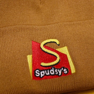 May include: A brown knitted beanie with a yellow and red embroidered logo that says "Spudsy's".