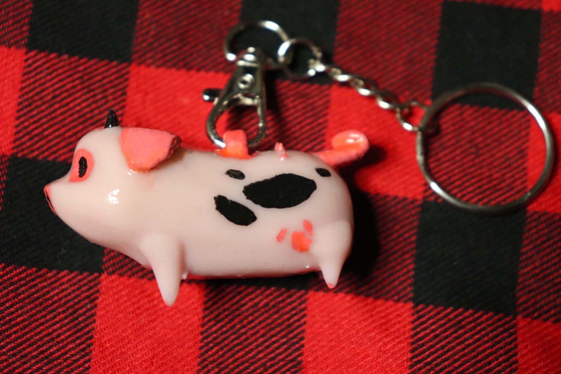 K&ouml;nnte beinhalten: Ein rosa Schweine-Schl&uuml;sselanh&auml;nger mit schwarzen Flecken und rosa Akzenten. Das Schwein ist an einem silbernen Schl&uuml;sselring befestigt.