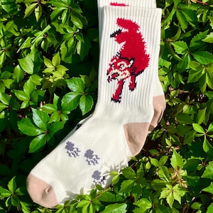 May include: A pair of off-white crew socks featuring a red fox graphic on the upper leg section, with tan-colored heels and toes. The foot area of the socks displays a pattern of small, dark gray paw prints arranged in a linear sequence.