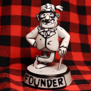 May include: A white figurine of a cartoon character wearing a red fez with a black outline. The character is wearing a white suit with black outlines and a black bow tie. The figurine is standing on a white base with the word "FOUNDER" written in black.